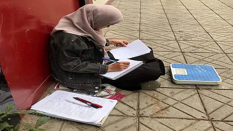 صورة مؤثرة لفتاة يمنية تذاكر في الشارع بجوار ميزان ترتزق منه